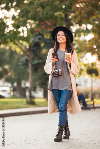 Modern asian woman holding mobile phone and coffee cup while walking in park outdoor