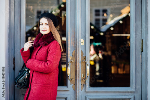 Stylish fashion girl in red coat and black beret, posing over old gray door of cozy cafe.Beauty girl in stylish coat. Fashion look.