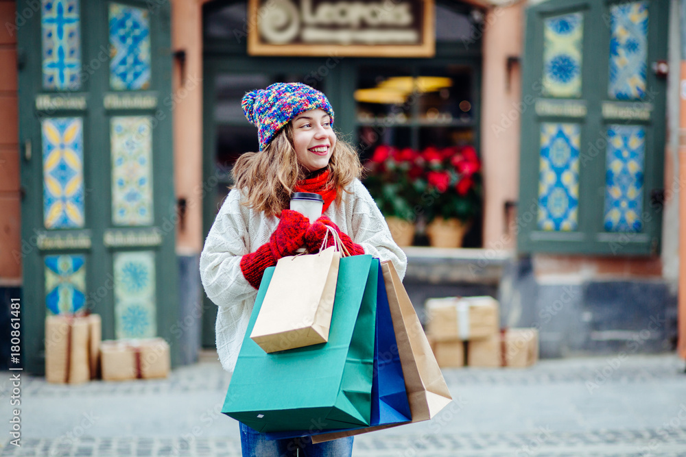 Young charming woman with shopping bags near shop with gift.Girl dressed in knitted hat, red scarf and mittens. Vacation, weekend, takeaway drinks, leisure concept.