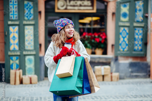 Young charming woman with shopping bags near shop with gift.Girl dressed in knitted hat, red scarf and mittens. Vacation, weekend, takeaway drinks, leisure concept.