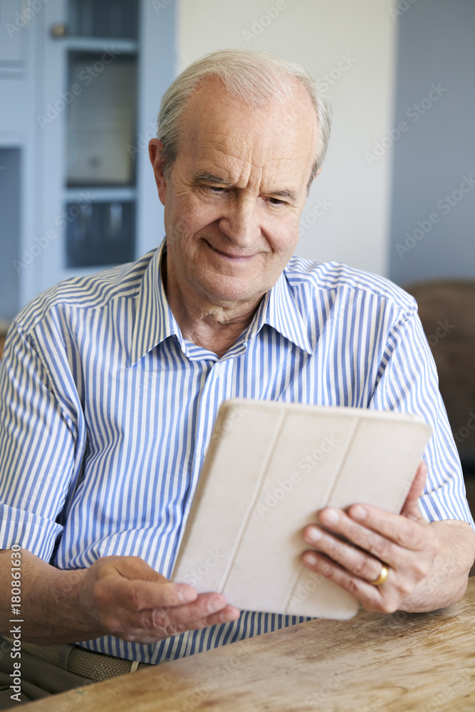 Senior Man Sitting At Home Using Digital Tablet At Table Senior Man Sitting At Home Using Digital Tablet At Table