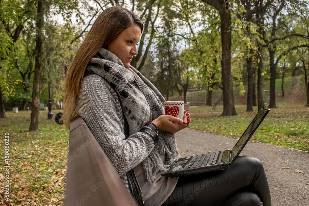 A young woman sitting on the bench watching in her laptop an holding a cup of tea A young woman sitting on the bench watching in her laptop an holding a cup of tea