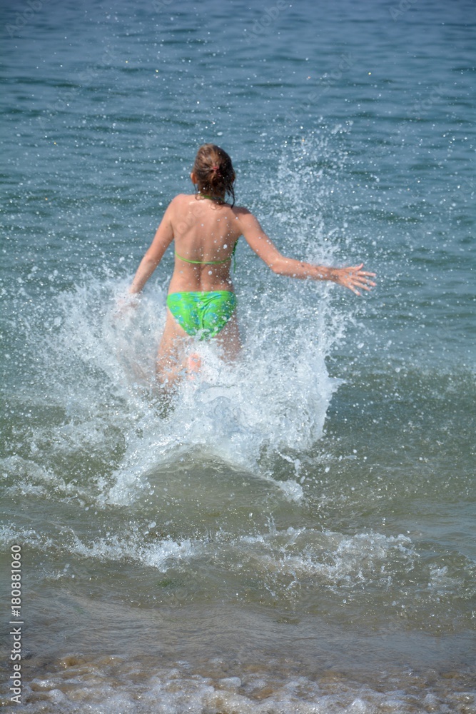 Mädchen im Bikini rennt ins Meer, Wasser spritzt nach oben