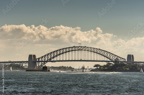 Sydney, Australia - March 26, 2017: Frontal view of black metalic Harbour Bridge including support towers on both sided seen off water under cloudscape. Denison Fort in bay and multiple small boats.