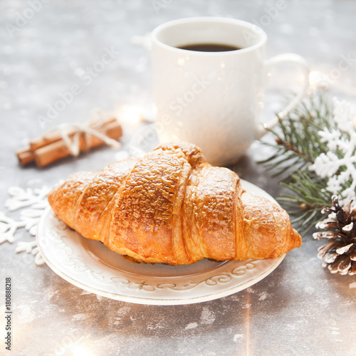Fresh croissant and cup of hot cofffee on cosy winter background with spuce and artificial lights.