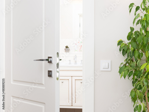 Ajar white door to the bathroom. Series switch on a light gray wall. Modern chrome door handle and lock