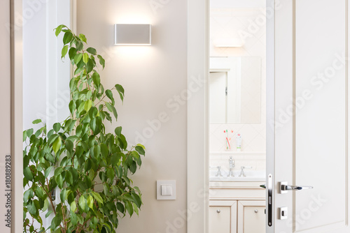 Ajar white door in the bright bathroom. Series switch and modern wall lamp on light gray wall. Chrome door handle and lock