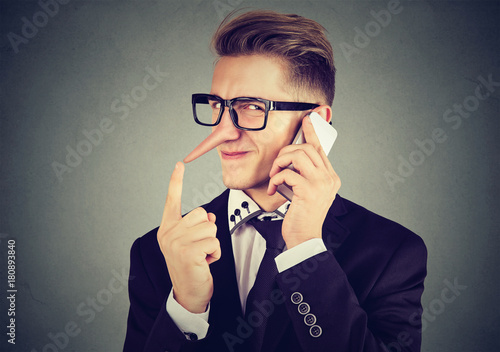 young sly man with long nose talking on mobile phone isolated on gray wall background. Liar concept. Human emotion feelings photo