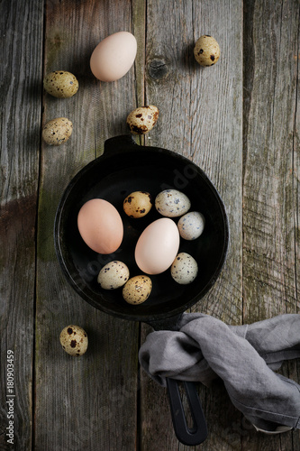 Whole quail and chicken eggs in a small cast-iron frying pan on the background of an old wooden texture. Selective focus. Top view with space