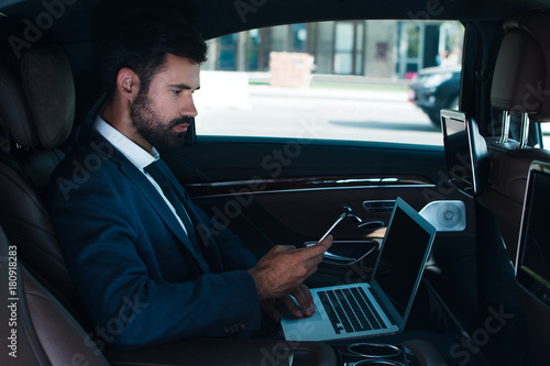 Checking emails on a go. Side view of handsome young man using laptop and using his smart phone while sitting in car © MARIIA