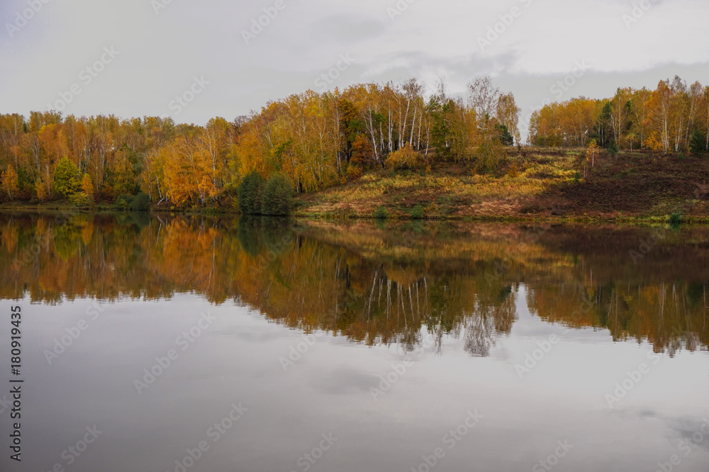 The bank of the river is reflected in the mirror calm water in the autumn cloudy day.