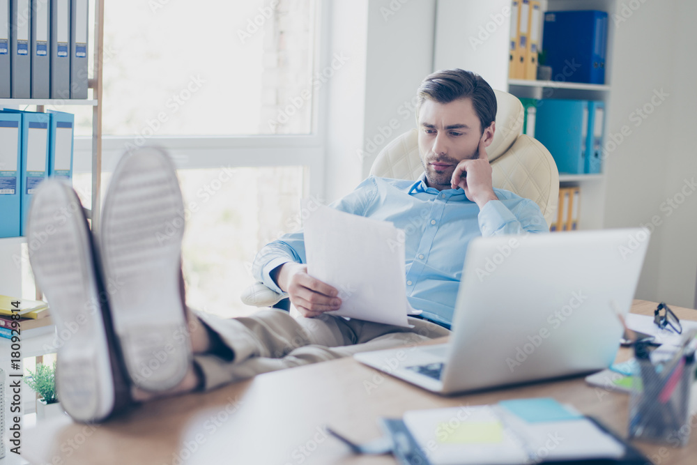 Young but experienced and confident investor is reading carefully the deposit agreement before signing it. He is relaxed and his feet are on the table Young but experienced and confident investor is reading carefully the deposit agreement before signing it. He is relaxed and his feet are on the table