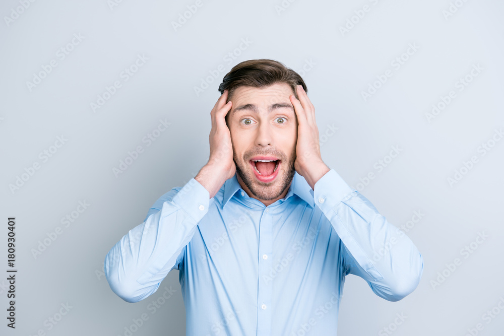 Close up portrait of happy, cute, attractive young man looking excited, surprised, full disbelief, opened his eyes and mouth holding his hands on the head isolated over grey background Close up portrait of happy, cute, attractive young man looking excited, surprised, full disbelief, opened his eyes and mouth holding his hands on the head isolated over grey background
