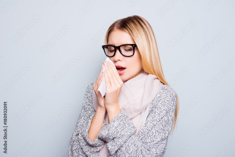 Concept of catching a cold in autumn. Close up portrait of young woman suffering from a cold. She is sneezing and using a paper tissue. She is wearing warm cardigan and scarf. Isolated on background Concept of catching a cold in autumn. Close up portrait of young woman suffering from a cold. She is sneezing and using a paper tissue. She is wearing warm cardigan and scarf. Isolated on background