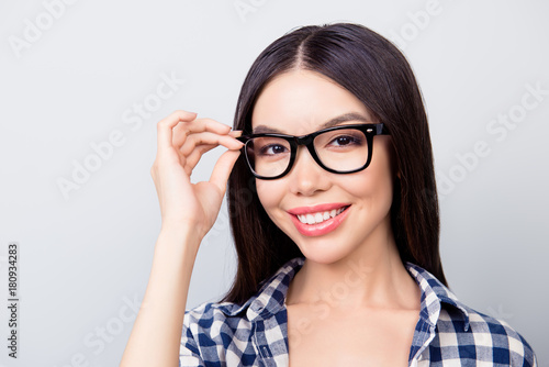 Close up portrait of charming smart lady with toothy smile, hazel eyes and beautiful face is touching her spectacles, dressed in casual checkered clothes isolated on grey background