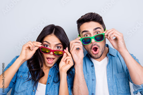 Concept of large sales and discount. Close up photo of two excited and wondered people with open mouths dressed in casual clothes, they are touching colorful glasses, isolated on grey background