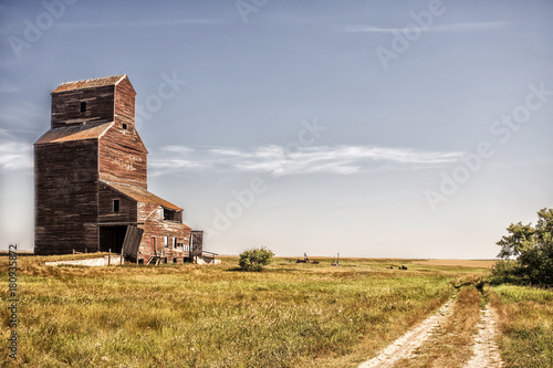 An abandoned old crumbling tall wooden grain storage elevator surrounded by agriculture crops in a desaturated rural countryside summer landscape