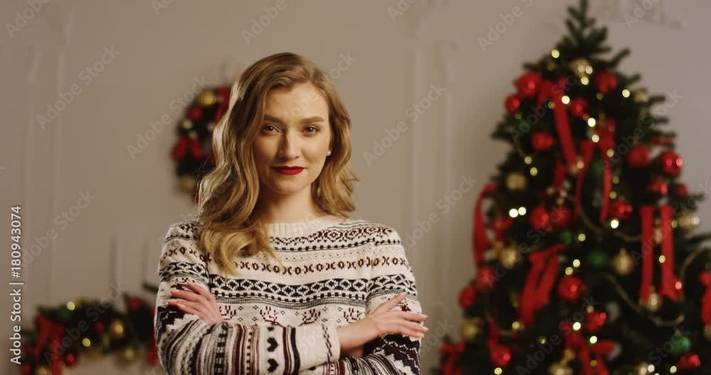 Portrait shot of young blonde pretty woman smiling sincerely and looking into the camera with crossed hands. The Christmas tree with red ribbons and lights background. Indoor. Close up Portrait shot of young blonde pretty woman smiling sincerely and looking into the camera with crossed hands. The Christmas tree with red ribbons and lights background. Indoor. Close up