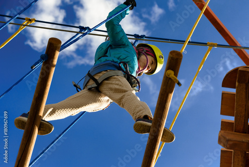 Happy girl in a yellow helmet and a blue jacket is playing in an adventure park holding ropes and climbing a wooden and rope staircase. Bottom view.