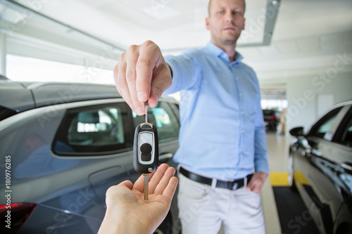 Male car dealer giving a car key to a female person hand. They are standing indoors in a showroom, with cars behind them