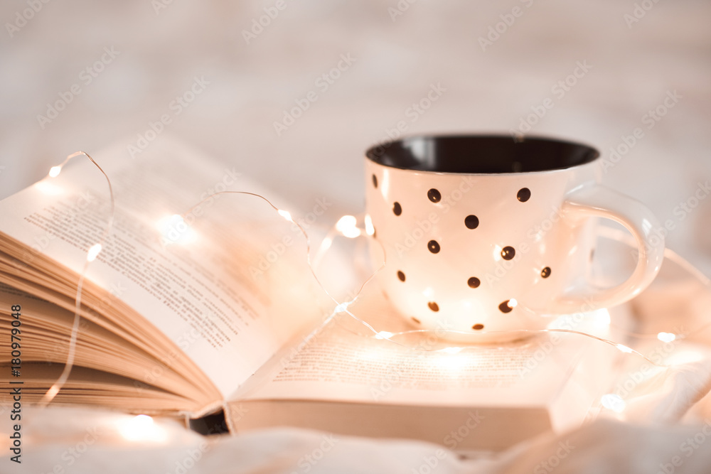 Cup of tea staying on open book with Christmas lights in bed closeup. Good morning. Breakfast time.