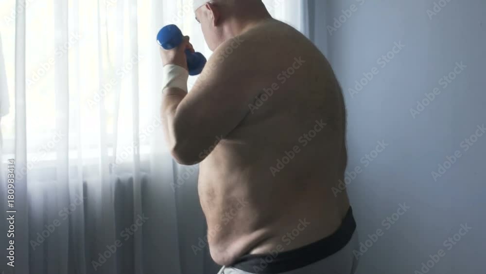 Fat man actively doing sports, raising dumbbells to lose weight, home exercising Fat man actively doing sports, raising dumbbells to lose weight, home exercising