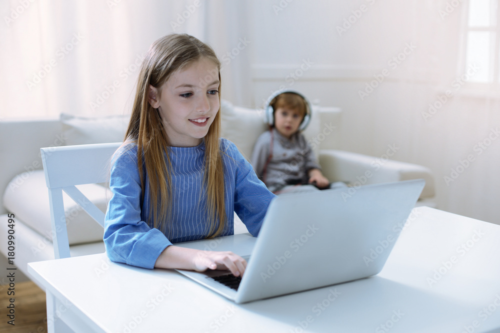 Progressive girl. Cheerful attentive smiling girl working at the laptop while her brother sitting on a sofa Progressive girl. Cheerful attentive smiling girl working at the laptop while her brother sitting on a sofa