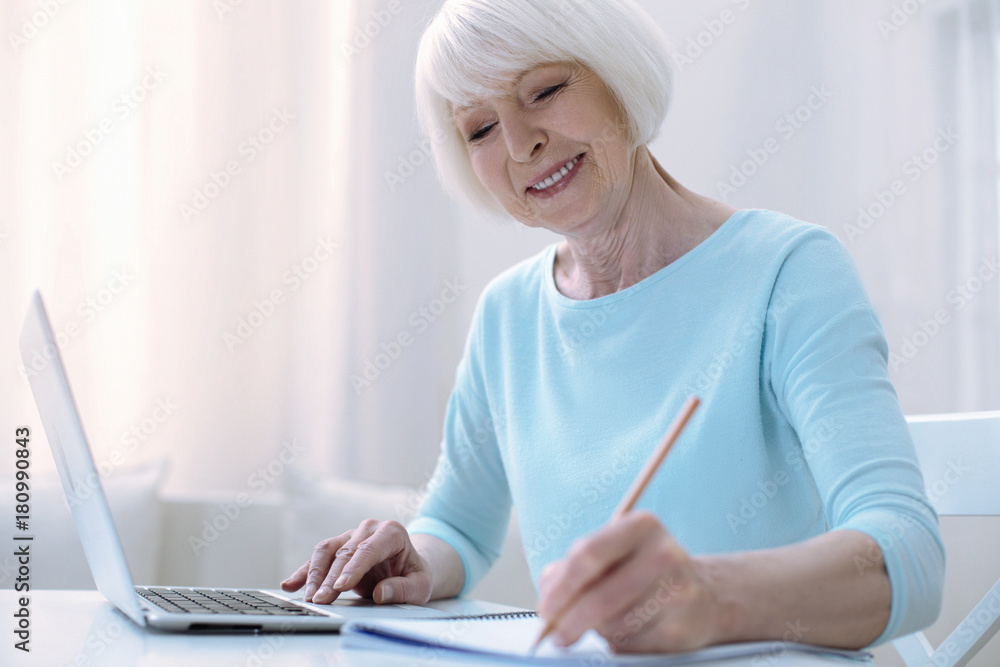Pleasant work. Nice positive old woman sitting in her room and working with a help of modern technologies Pleasant work. Nice positive old woman sitting in her room and working with a help of modern technologies