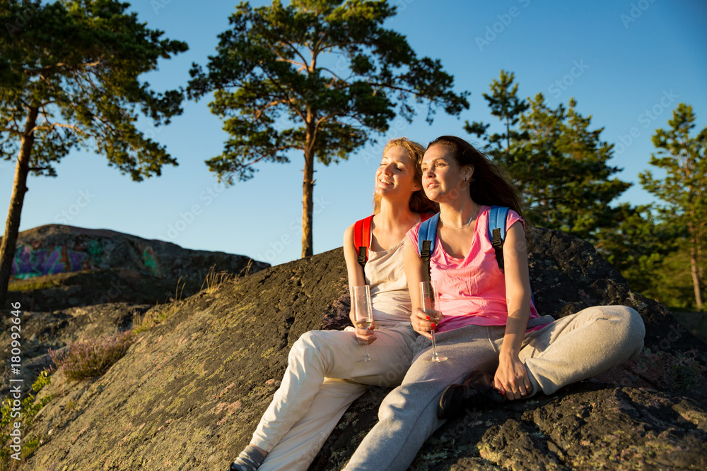 Two women hiking on top of the rock. Sitting, drinking champagne , celebrating, enjoying scenic view in Finland. Girl power concept. 