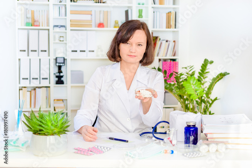 Female doctor gives the patient medicines or pills at workplace in office with the bookshelves in the background und various medications, pills and bandages on the table.