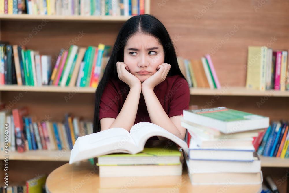 Asian woman student boring reading book at library with a lot of books in university. Asian student disheartened reading book for examination.. Asian woman student boring reading book at library with a lot of books in university. Asian student disheartened reading book for examination..