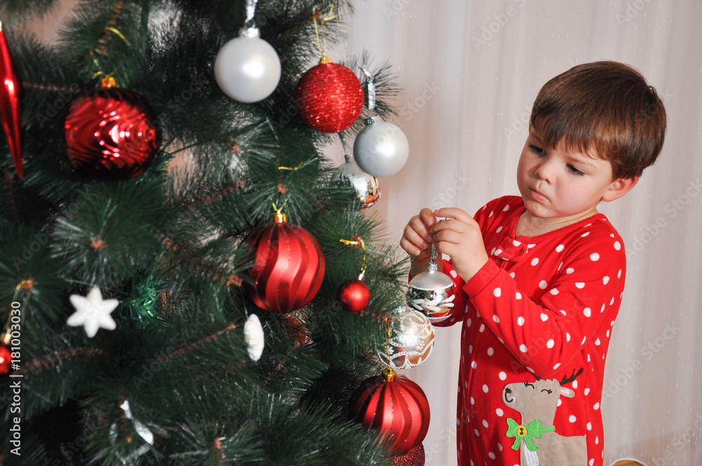 Boy in New Year's pajama decorating Christmas tree. Child putting decorations on Christmas tree Boy in New Year's pajama decorating Christmas tree. Child putting decorations on Christmas tree