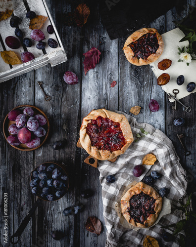 plum crostata pies on gray wooden table with textile, fresh plums in plate and in pan, old keys and dry leaves