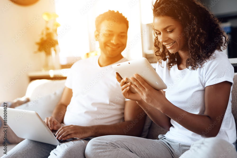 Black man and woman are sitting on the couch. A man is working on a laptop, a woman is reading something on a tablet. Black man and woman are sitting on the couch. A man is working on a laptop, a woman is reading something on a tablet.