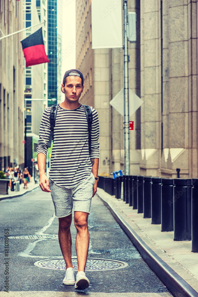 Man summer casual street fashion in New York. Wearing black tank top,  shorts, white sneakers, cap worn backward, a young guy stands against wall,  reading messages on cell phone. Filtered effect.. Stock