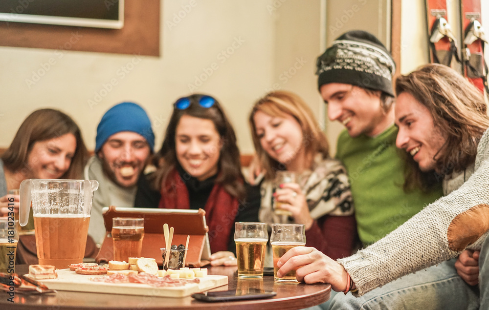 Happy friends watching videos and drinking beer at chalet pub in mountains - Young people having fun in winter vacation - Friendship concept - Focus on right man left hand - Warm filter