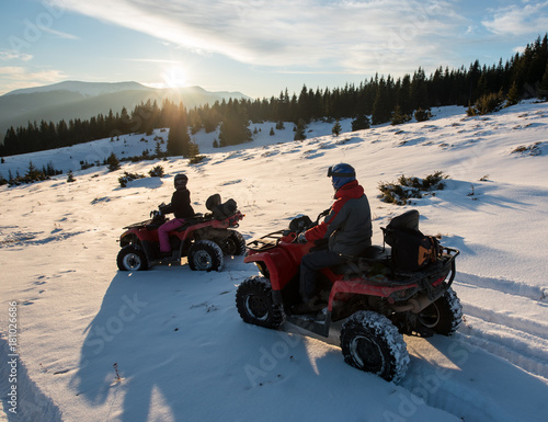 Young couple riders sitting on four-wheelers ATV bikes on snow, enjoying sunset in the the mountains in winter