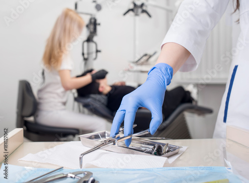 Close-up view of assistant's hands with blue gloves working with dental equipment, on the blurred background female dentist is treating patient in dental office. Dentistry