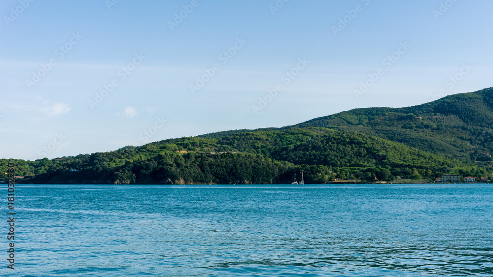 The cliff covered by trees and green bush and sea with few boats on water