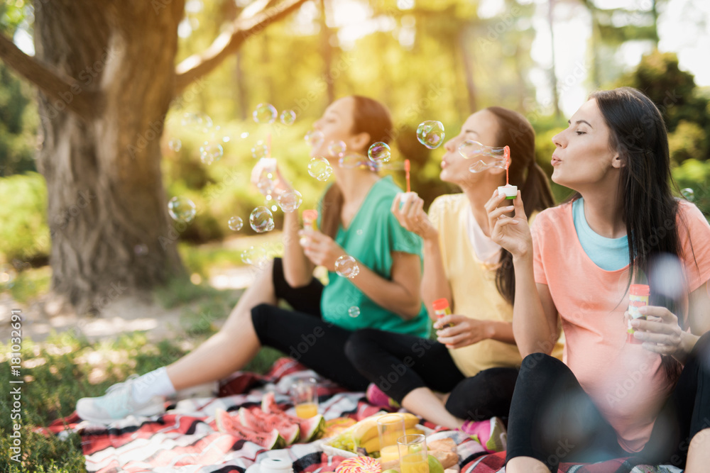 Fototapeta premium Three pregnant women sit in a park on a picnic rug and blow bubbles