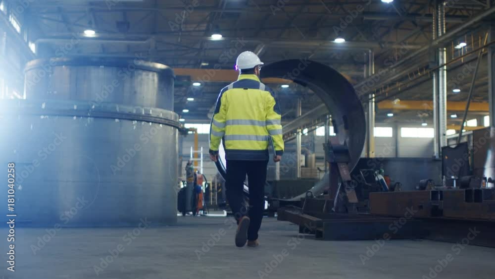 Industrial Engineer in Hard Hat Wearing Safety Jacket Walks Through Heavy Industry Manufacturing Factory with Various Metalworking Processes. Industrial Engineer in Hard Hat Wearing Safety Jacket Walks Through Heavy Industry Manufacturing Factory with Various Metalworking Processes.