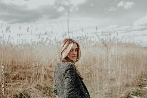 Pretty and young woman with long blonde hair dressed in wool coat looking at camera over her shoulder and smiling. Background bulrush. Autumn. Outdoor. Medium shot