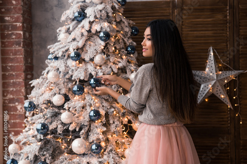 christmas , winter holidays and people concept - young asian woman decorating christmas tree with ball at home