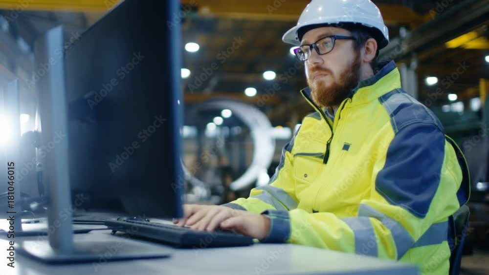 Industrial Engineer Works at Workspace on a Personal Computer.  He Wears Hard Hat and Safety Jacket and Works in the Main Workshop of the Heavy Industry Manufacturing Factory.