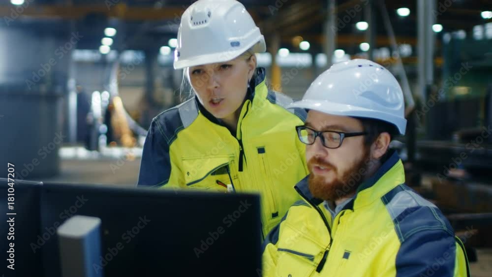 Male Industrial Engineer Works on the Personal Computer while Female Factory Worker Talks about Project Details. They Work in Heavy Industry Manufacturing Facility Male Industrial Engineer Works on the Personal Computer while Female Factory Worker Talks about Project Details. They Work in Heavy Industry Manufacturing Facility