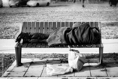 Foto Homeless refugee man in black coat sleeps on the bench on the street black and w