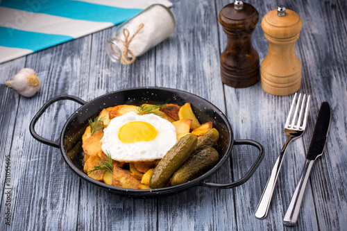 Fried eggs and potatoes in a frying pan with pickles on wooden rustic background.