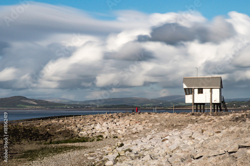 An elevated hut looks out over a pebble beach towards the Morecambe Bay, Lancashire with rolling clouds above. This is a panorama stitch of 5 images.