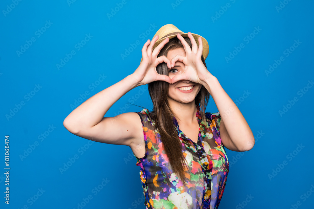 Beautiful young woman in sun hat is showing heart shape hand sign on face and smiling on blue background Beautiful young woman in sun hat is showing heart shape hand sign on face and smiling on blue background