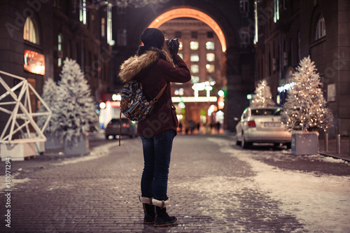 Hipster girl walking through the night city street taking photos of buildings and Christmas trees